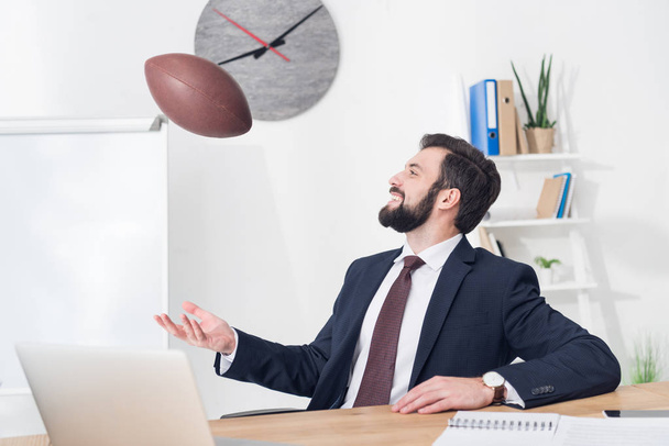 портрет бизнесмена, бросающего регбийный мяч на рабочем месте с ноутбуком в офисе
 - Фото, изображение