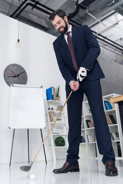 низкий угол обзора улыбающегося бизнесмена в костюме, играющего в гольф в офисе
 - Фото, изображение