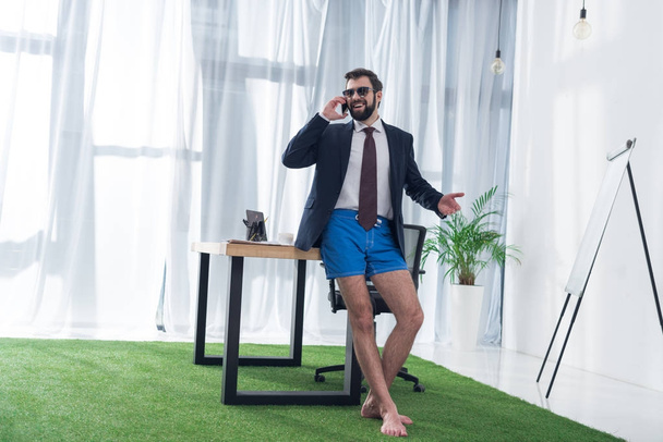 businessman in jacket and shorts talking on smartphone at workplace in office - Fotó, kép