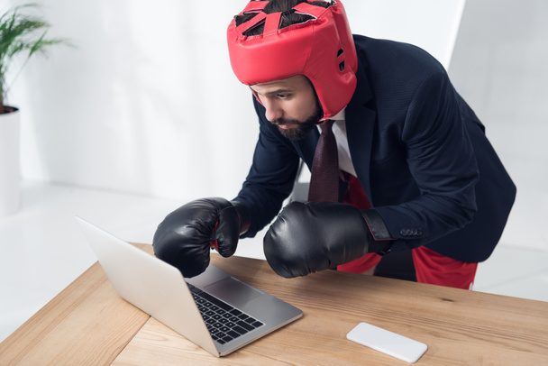 бизнесмен в боксёрских перчатках и шлеме печатает на ноутбуке на рабочем месте в офисе
 - Фото, изображение