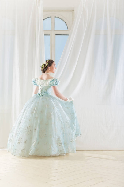 Κομψή γυναίκα σε ένα υψηλό παράθυρο στο ιστορικό φόρεμα, ισιώνει τις κουρτίνες, και περιμένει τους φίλους για χορό να χορέψει - Φωτογραφία, εικόνα