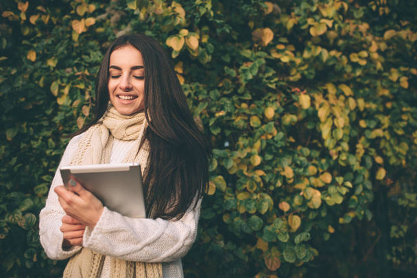 Krásná mladá brunetka sedí na spadané podzimní listí v parku, čtení e knihy v tabletovém počítači. - Fotografie, Obrázek