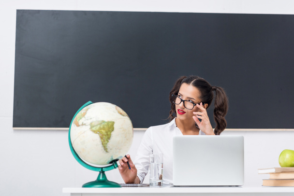 promyšlené sexy učitel sedí na pracovišti s globe a notebooku v učebně - Fotografie, Obrázek