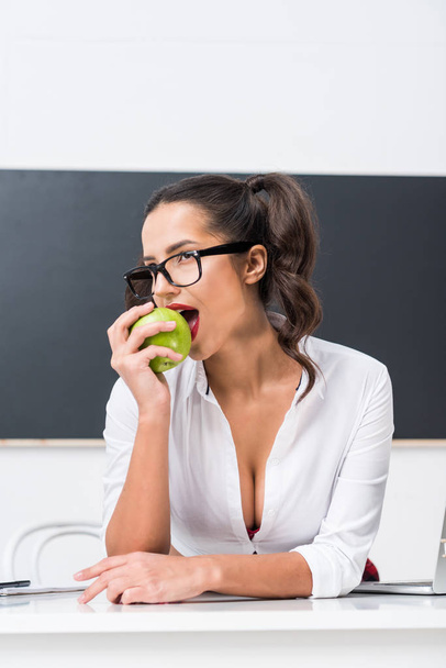 Nuori seksikäs opettaja syö omenaa työpöydällä luokassa
 - Valokuva, kuva