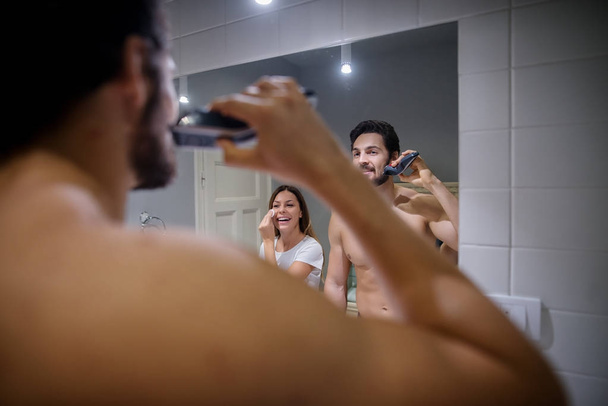 Szczęśliwa młoda miłość para razem w łazience robi rano codziennej pielęgnacji. Człowiek jest golenie z golarka elektryczna - Zdjęcie, obraz
