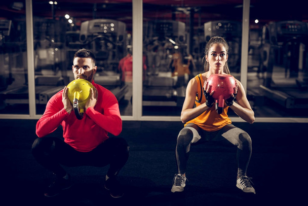 strong young sporty couple in sportswear doing squats with kettlebells in gym - Photo, Image