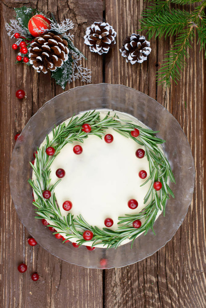 Homemade cake for Christmas with cream cheese frosting, decorated with rosemary and berries on a wooden table - Foto, afbeelding