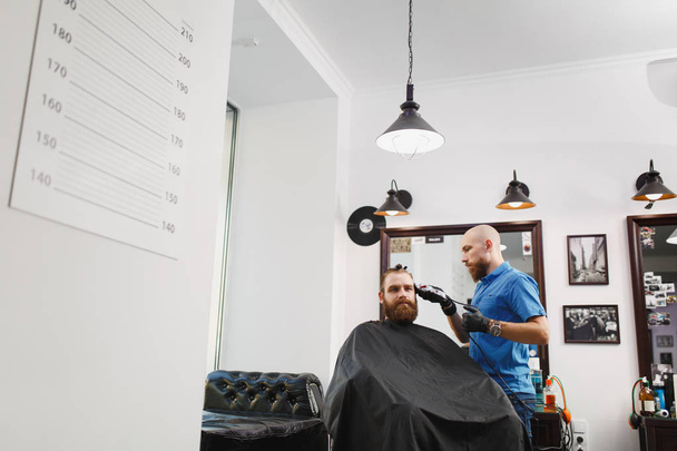 Male professional hairdresser serving client by clipper. Ginger handsome brutal stylish young man with thick big beard and short hair getting trendy haircut in black cape. Light white barber shop room - Photo, Image