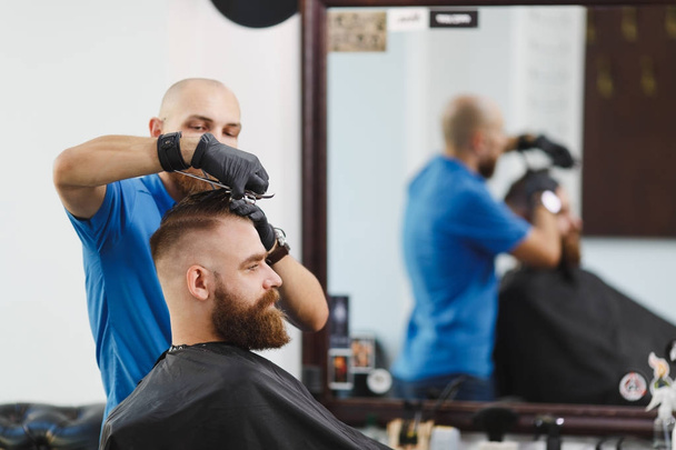 Male professional hairdresser serving client by scissors. Ginger handsome brutal stylish young man with thick big beard, short hair getting trendy haircut in black cape. Light white barber shop room. - 写真・画像