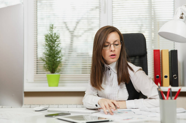 Poważne i pochłonięty biznes brązowy włosy kobieta w garnitur i okulary siedzi przy biurku z tabletu, praca przy komputerze z nowoczesnego monitora z dokumentów w biurze światła, patrząc na bok - Zdjęcie, obraz