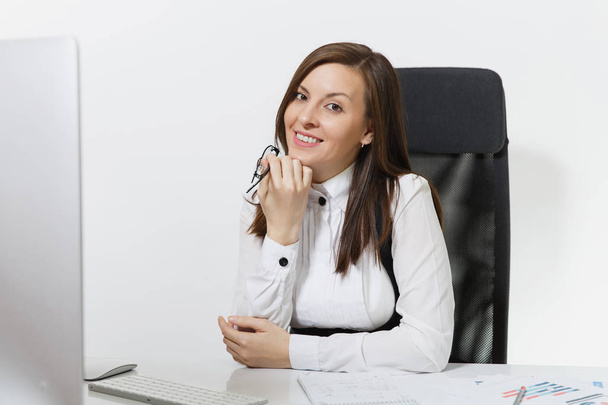 Na białym tle piękny uśmiechający się brązowe włosy kobieta biznesu w garnitur i okulary, siedząc przy biurku, praca przy komputerze z nowoczesnego monitora z dokumentów w biurze światła, patrząc na kamery - Zdjęcie, obraz