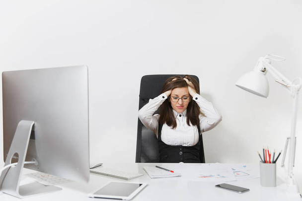 Красивая уставшая смущенная и напряженная каштановые волосы деловая женщина в костюме и очках сидит за столом, работая за современным компьютером с документами в светлом офисе на белом фоне
 - Фото, изображение