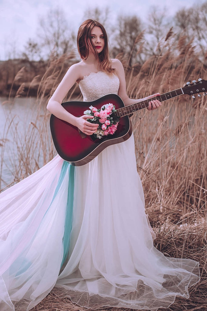 Prachtig romantisch europees meisje met gitaar met bloemen erin, poserend buiten. Concept van muziek en natuur. Voorjaarstijd. - Foto, afbeelding