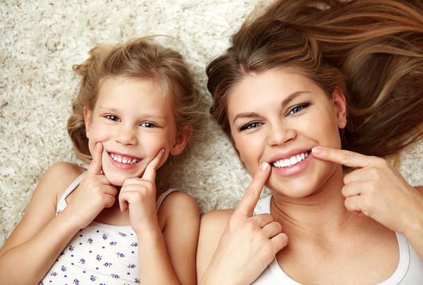 Portrait of beautiful daughter with mom showing their healthy white smile lying on carpet at home. - Foto, Imagem