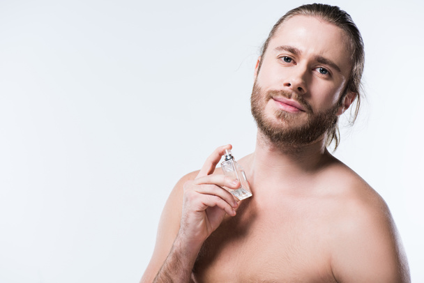 Bearded young man looking at camera while holding glass bottle with perfumes against his neck, isolated on white   - Foto, Bild