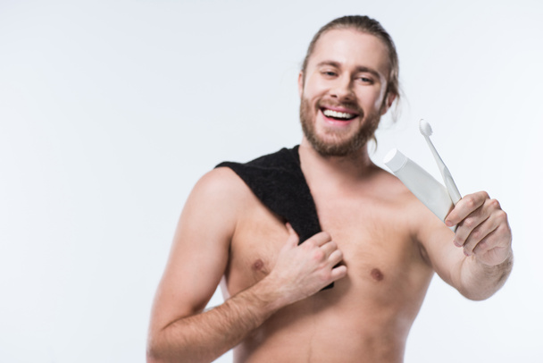 Χαμογελαστός νεαρός άνδρας με το μαύρο πετσέτα στον ώμο κρατώντας οδοντόκρεμα και οδοντόβουρτσα στο χέρι, απομονώνονται σε λευκό - Φωτογραφία, εικόνα