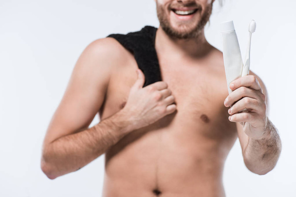 Imagen recortada de hombre barbudo caucásico con toalla negra en el hombro sosteniendo pasta de dientes y cepillo de dientes, aislado en blanco
 - Foto, Imagen