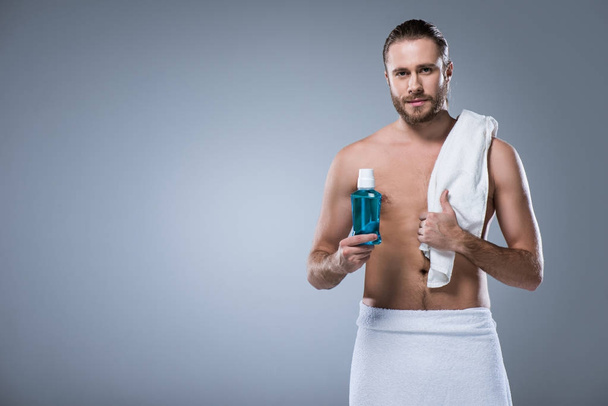 man with bath towel on shoulder holding bottle with tooth rinse in hand, isolated on gray  - Photo, Image