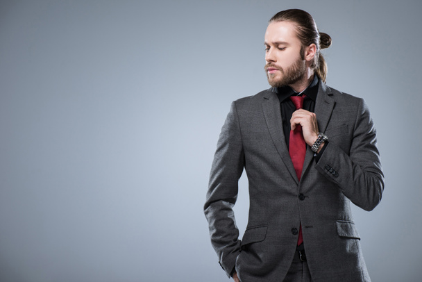 Joven hombre de negocios con los ojos cerrados tocando su corbata roja mientras otra mano en el bolsillo de la chaqueta, aislado en gris
 - Foto, Imagen