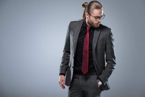 Hombre de negocios morena de pelo largo en gafas mirando hacia abajo con la mano en el bolsillo, aislado en gris
 - Foto, imagen