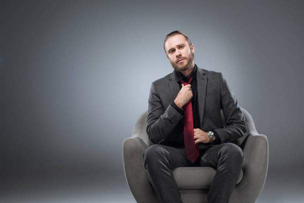 Hombre de negocios serio sentado en sillón y enderezando la corbata, mientras mira a la cámara, aislado en gris
 - Foto, imagen