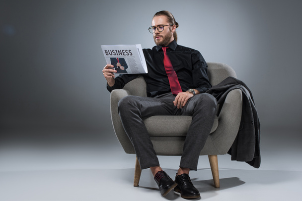 Caucasian bearded businessman in glasses sitting in armchair and reading newspaper, isolated on gray    - Fotografie, Obrázek