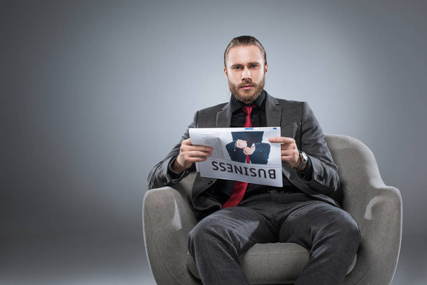 Homem de negócios barbudo caucasiano sentado em poltrona e com jornal nas mãos enquanto olha para a câmera, isolado em cinza
    - Foto, Imagem