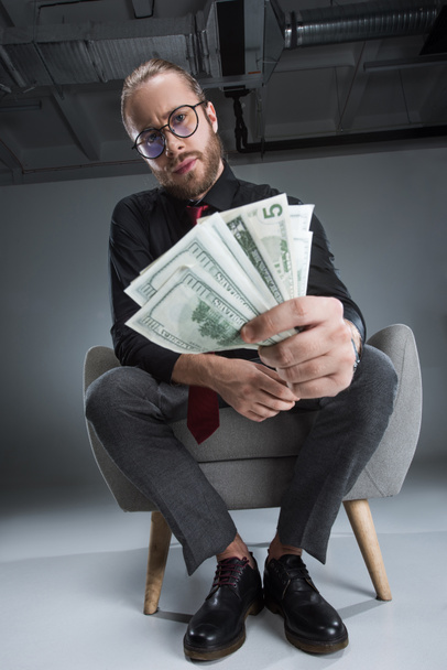 Vakava liikemies lasit istuu nojatuolissa rahaa käsissä ja katselee kameraa
 - Valokuva, kuva