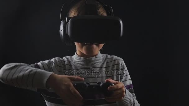 Boy uses VR-headset display with headphones and joystick for virtual reality game at black background - Footage, Video