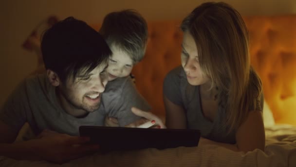 Happy family with little son using tablet computer and talking lying in bed at home before sleeping - Materiał filmowy, wideo