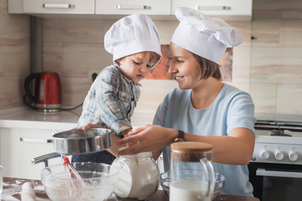 ευτυχισμένη μητέρα και αξιολάτρευτο παιδί στο chef καπέλα ζύμη στο ακατάστατο τραπέζι στην κουζίνα - Φωτογραφία, εικόνα