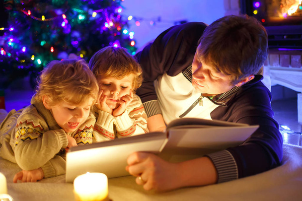 Ojciec i dwóch małych chłopców malucha czytanie książki przez komin, świece i kominkiem. - Zdjęcie, obraz