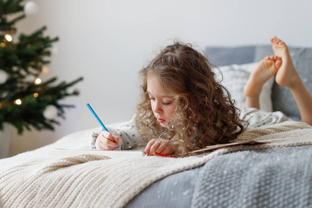 Portrait of adorable curly girl spends free time on bed, makes Christmas card for parents, writes wishes and draws pictures, has concentrated look, being obidient kid. New Year and children concept. - Foto, imagen