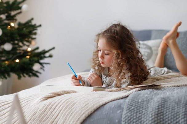 Indoor shot of attentive pretty small girl writes letter to Santa Claus before Christmas, thinks what present she expects, wants all dreams and wishes come true, lies on comfortable bed indoor. - Foto, Imagen