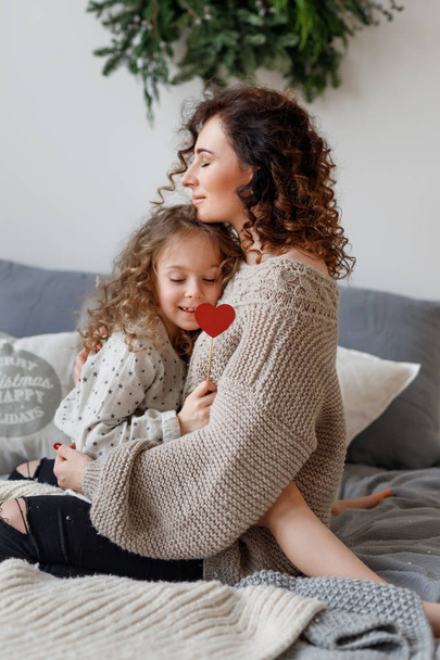 Vertical shot of affectionate young mother and her daugher embrace each other and express love, sit on comfortable bed, close eyes with enjoyment, being relaxed and pleased. Parenthood concept. - Zdjęcie, obraz
