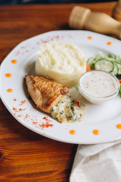 Closeup view of baked chicken breast stuffed with cheese supplemented with creamed potatoes, sour cream sauce and sliced cucumbers on salad - Photo, Image