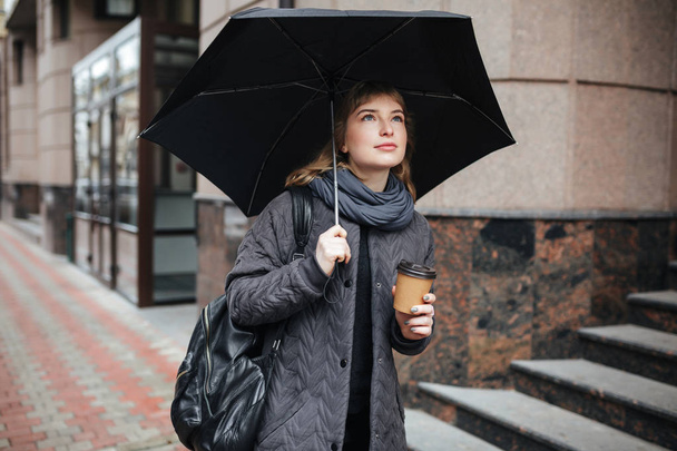 Portrait of young cute lady standing on street with black umbrella and coffee in hands - Foto, immagini