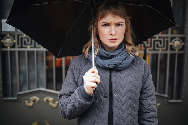 Portrait of upset lady standing on street with black umbrella and  sadly looking in camera isolated - Foto, Imagem