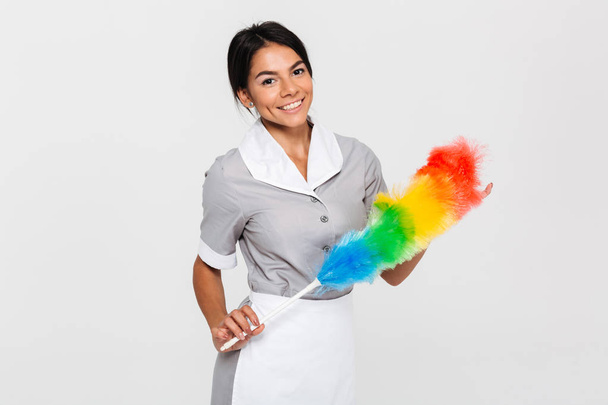Cheerful brunette housekeeper in uniform holding colorful duster - Foto, Imagem