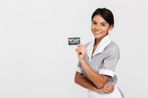 fröhliche Brünette Dienstmädchen in Uniform mit Kreditkarte und Blick in - Foto, Bild