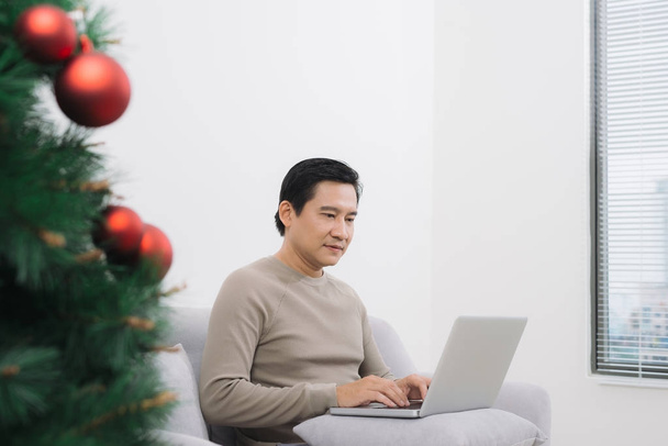 Hombre con portátil mientras está sentado en el sofá en el árbol de Navidad
 - Foto, Imagen