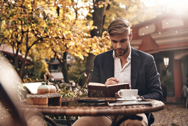 Good looking young man in smart casual wear looking at his personal organizer while sitting in restaurant outdoors - Foto, Imagen