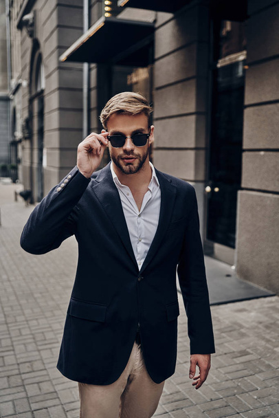 Handsome young man in smart casual wear adjusting eyewear while walking in city street - Foto, Bild