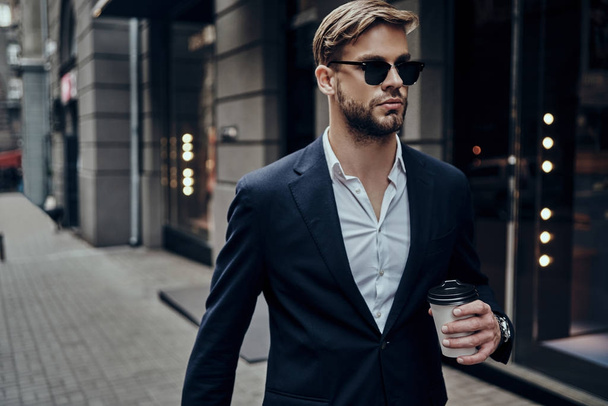 Przystojny młody biznesmen w codzienny nosić, niosąc kubek jednorazowy, podczas spaceru w ulicy miasta - Zdjęcie, obraz