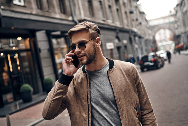 Handsome young man in casual wear talking on smart phone while standing outdoors - Fotó, kép