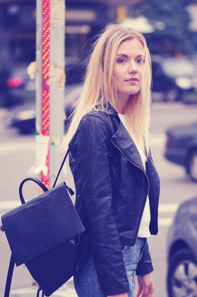 Pretty blonde girl posing outdoors. The girl is dressed in a black leather jacket and a white sweater. On his shoulder is a black bag. Cars on a blurry background. - Foto, immagini