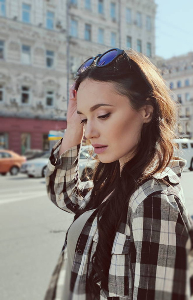 Portrait of a beautiful brunette woman in a plaid shirt. Urban style of clothes. Sunglasses on the head. Reflection of the sky in glasses glasses. - 写真・画像