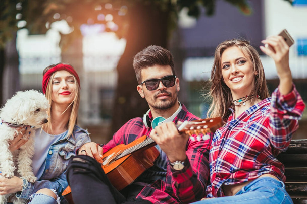 Happy νεαρό φίλοι διασκεδάζουν παίζοντας κιθάρα και λήψη selfie φωτογραφία στο πάρκο  - Φωτογραφία, εικόνα