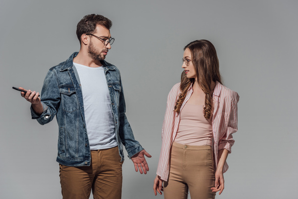 Stilvoller junger Mann mit Brille, Smartphone in der Hand und Freundin isoliert auf grau anschauend - Foto, Bild