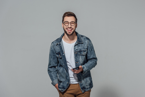 cheerful young man holding smartphone and smiling at camera isolated on grey - Photo, Image
